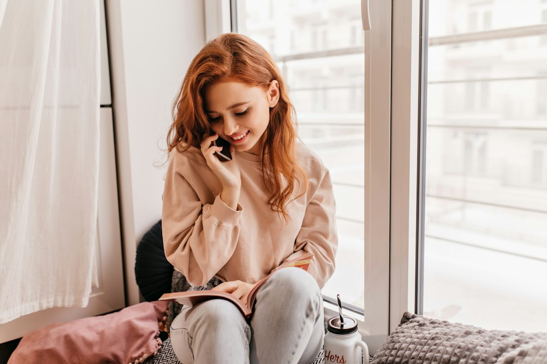 窓辺に座ってスマートフォンで通話する女性とひざの上のノート