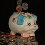暗い背景に置かれた陶器の豚の貯金箱と、上から落ちてくる硬貨