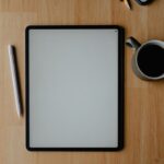 白いタブレット端末とApple Pencil、コーヒーカップが置かれた木製デスク