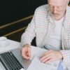ノートパソコンの前で書類を確認しながら考え込む男性