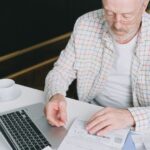 ノートパソコンの前で書類を確認しながら考え込む男性