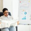 オフィスでグラフが貼られたホワイトボードの前で新聞を読む女性