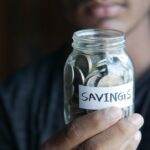 SAVINGSと書かれたラベルを貼った硬貨入りのガラス瓶を手に持つ様子