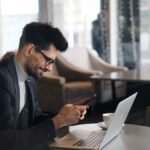 カフェでノートパソコンを前にスマートフォンを操作するビジネスマン