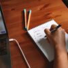 ノートに手書きでメモを取る人物の手元とノートパソコン