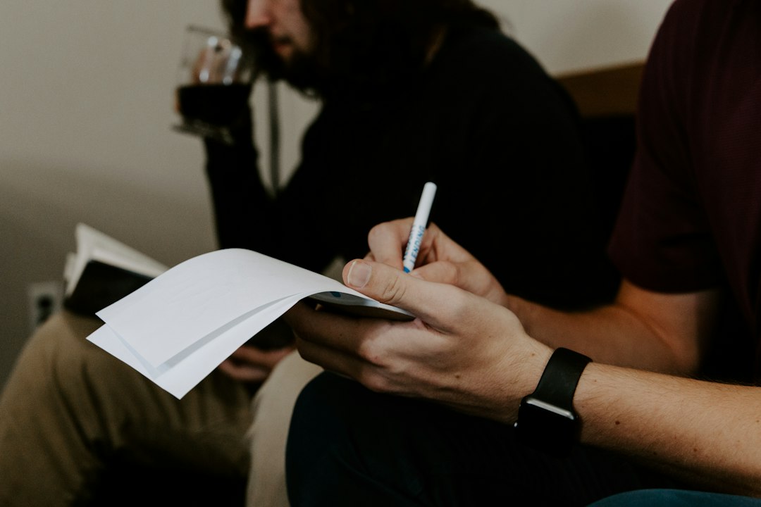 ノートにペンで書き込む手元と、隣で飲み物を手にする人物